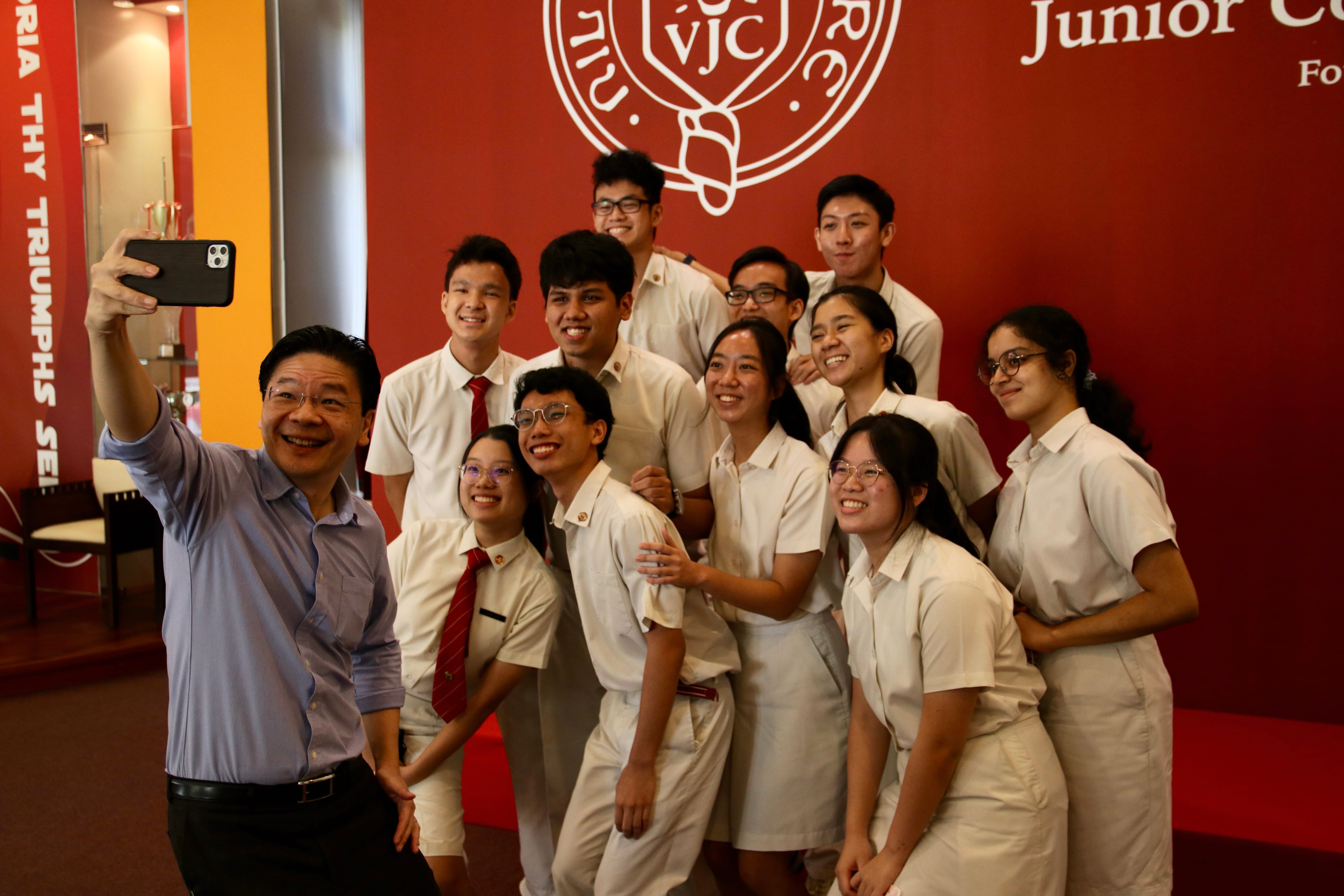 A man takes a selfie with a group of smiling students in white uniforms in front of a red VJC Junior College wall.